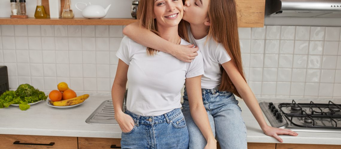 Loving teenage girl kissing smiling mother on cheek while siting on counter in kitchen